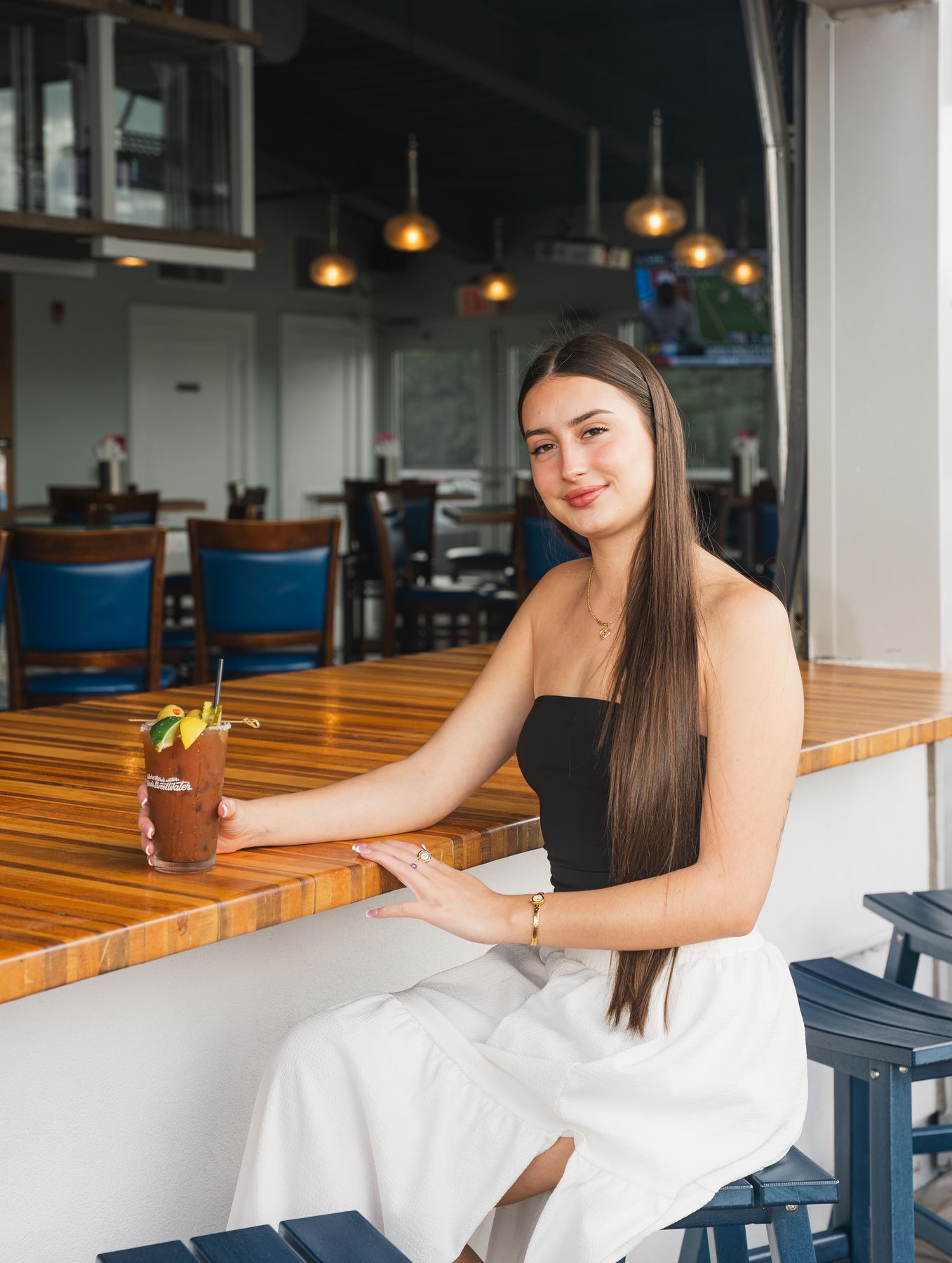 Woman in a black top seated at a wooden bar holding a garnished Bloody Mary.