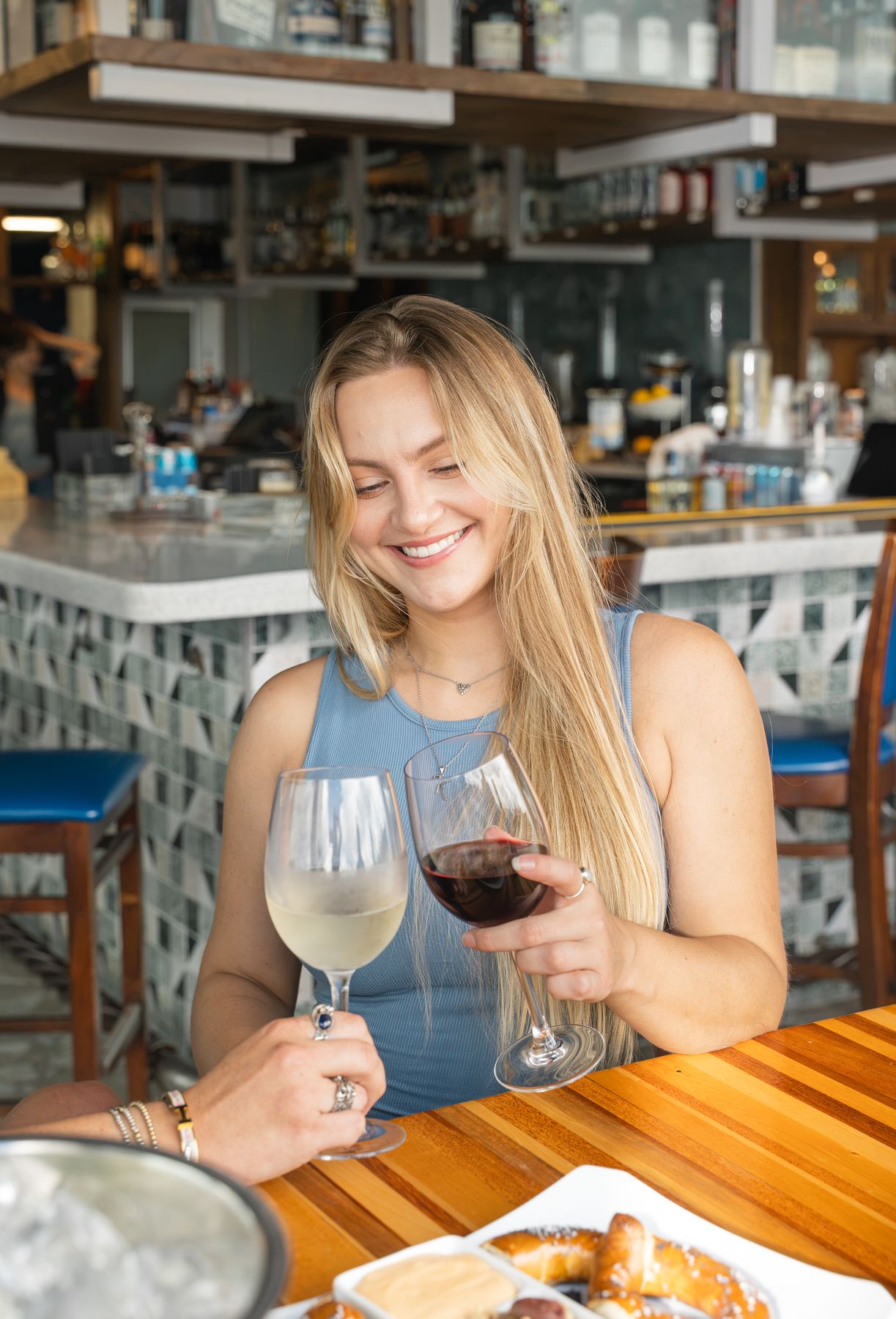Smiling woman toasting a glass of red wine with a friend's white wine at a bright bar.